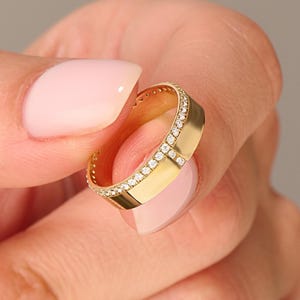 May include: A gold ring with a band of small, sparkling diamonds. The ring has a flat, rectangular design with a polished finish. The ring is held between two fingers, showcasing its elegant and modern style.