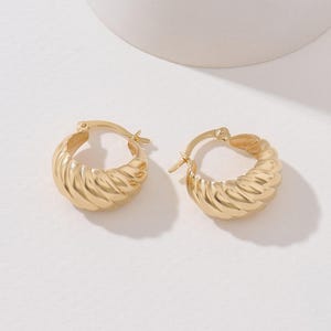 May include: A pair of gold-toned hoop earrings featuring a thick, rounded, and twisted croissant-like texture. Each earring is designed with a hinged post closure mechanism, showcasing a polished metallic finish that reflects light across the ridges.