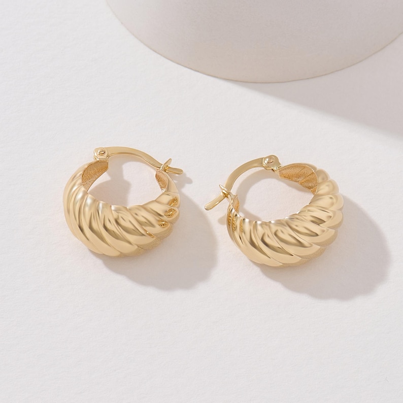 May include: A pair of gold-toned hoop earrings featuring a thick, rounded, and twisted croissant-like texture. Each earring is designed with a hinged post closure mechanism, showcasing a polished metallic finish that reflects light across the ridges.