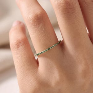 May include: A thin gold band ring featuring a series of small, round, vibrant green gemstones set along the top half of the band. The ring has a delicate, minimalist profile with a polished metal finish and evenly spaced green stones.