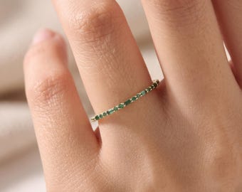 Puede incluir: Un anillo de banda fina de oro que presenta una serie de pequeñas piedras preciosas redondas de color verde vibrante engastadas a lo largo de la mitad superior de la banda. El anillo tiene un perfil delicado con un acabado de metal pulido.
