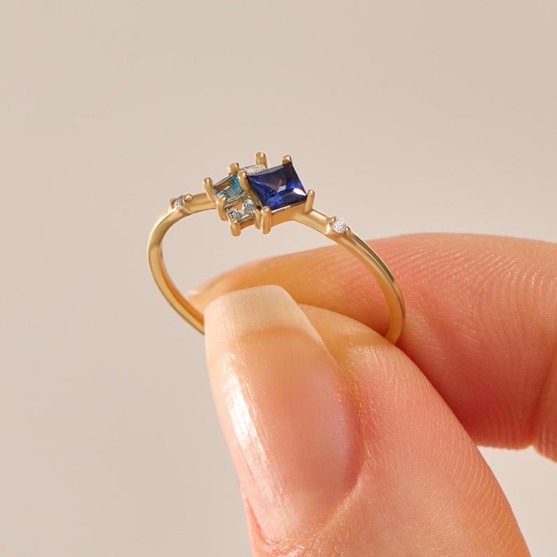 May include: A gold ring featuring a square, deep blue gemstone and two smaller, light blue and clear gemstones. The ring has a delicate band with small, clear accent stones. The ring is held by a person.