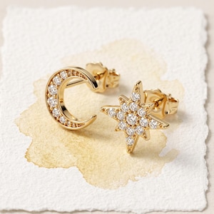 May include: A pair of gold stud earrings featuring a crescent moon shape and a multi-pointed star shape. Both earrings are set with small, clear, round-cut stones along their front surfaces and include standard post-style backings.
