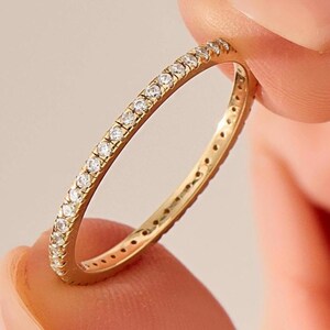 May include: A gold band ring with a continuous row of small, clear gemstones. The ring is held between two fingers, showcasing its delicate design and sparkling details. The ring's band is a warm gold color.
