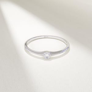 May include: A delicate silver ring with a single, round, clear gemstone set in the center. The ring band is thin and polished, reflecting light. The ring is set against a white background.