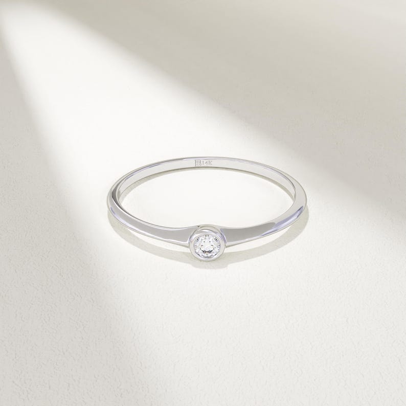 May include: A delicate silver ring with a single, round, clear gemstone set in the center. The ring band is thin and polished, reflecting light. The ring is set against a white background.