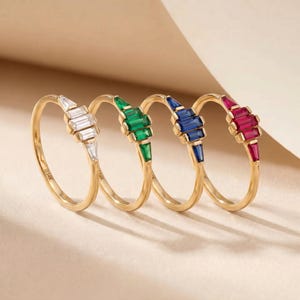 Op de afbeelding: Vier gouden ringen met kleurrijke edelsteenaccenten. Elke ring heeft een andere edelsteen: wit, groen, blauw en rood. De edelstenen zijn rechthoekig en in een lineair patroon gerangschikt, wat een moderne uitstraling creëert.