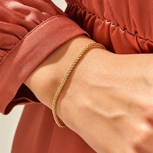 May include: A thin, gold-toned wheat chain bracelet with a woven, textured link design. The bracelet is worn around the wrist, showcasing a continuous, flexible metal construction with a polished finish that reflects light across its surface.