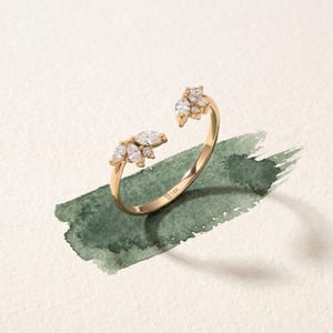 Könnte beinhalten: Ein offener Ring aus 14K Gold mit zwei Clustern aus klaren Steinen im Marquise- und Rundschliff an beiden Enden. Das Band besteht aus poliertem Gold und die Innenseite des Rings ist deutlich mit dem Text 14K für den Feingehalt geprägt.