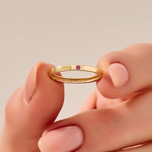 Könnte beinhalten: Ein goldener Bandring mit einem kleinen, runden, roten Edelstein in der Mitte. Der Ring wird zwischen zwei Fingern gehalten, was sein zartes Design und seine polierte Oberfläche hervorhebt. Der Ring ist etwa 0,25 cm breit.