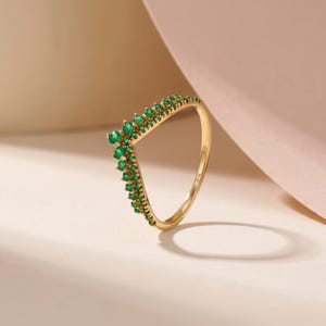 May include: A gold-colored ring with a V-shaped design, adorned with small, vibrant green gemstones. The ring's band is smooth and polished, and the gemstones are set along the V-shape, creating a delicate and elegant appearance. The ring is displayed on a light-colored surface.