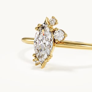 Op de afbeelding: Een gouden ring met een centrale diamant in marquise-slijpvorm, geflankeerd door kleinere diamanten in ronde slijpvorm in een clusteropstelling. De ringband is dun en gepolijst en heeft een glanzende geelgouden afwerking die contrasteert met de heldere stenen.