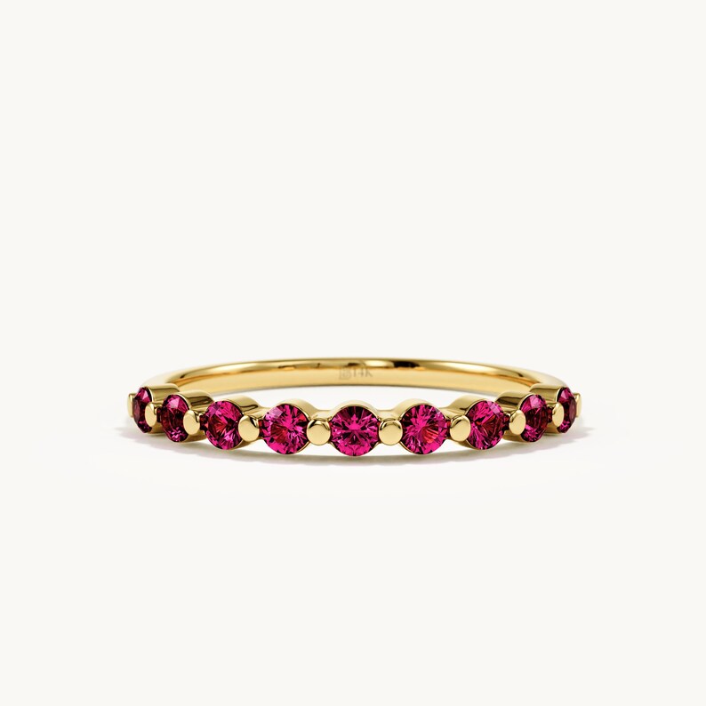 May include: A gold band ring featuring a row of seven round-cut vibrant pink gemstones. Each gemstone is set in a circular gold setting, alternating with small gold spheres along the band. The metal has a polished finish and a thin, delicate profile.