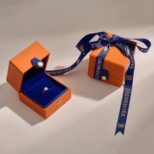 Puede incluir: Una caja de anillos naranja abierta con un interior azul muestra un anillo de diamantes. Una caja naranja cerrada est&aacute; atada con una cinta azul impresa con "BRENNMORE". Las cajas tienen cierres y detalles dorados. El anillo es dorado.