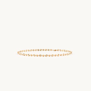 May include: A gold chain necklace featuring a series of small, circular, textured discs linked together in a continuous row. The metallic surface of each disc reflects light, creating a shimmering effect along the length of the delicate jewelry piece.