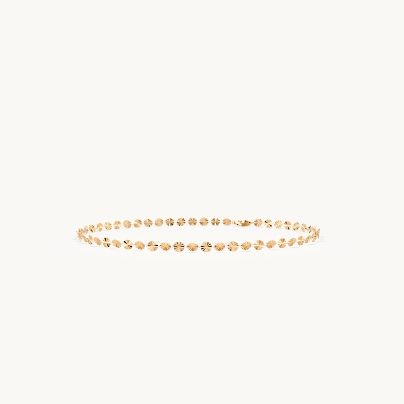May include: A gold chain necklace featuring a series of small, circular, textured discs linked together in a continuous row. The metallic surface of each disc reflects light, creating a shimmering effect along the length of the delicate jewelry piece.