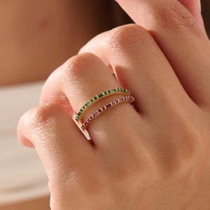 Op de afbeelding: Twee gouden ringen, &eacute;&eacute;n met groene edelstenen en de andere met roze edelstenen, zijn op een vinger gestapeld. De ringen hebben een delicaat, minimalistisch ontwerp, met de edelstenen dicht bij elkaar.