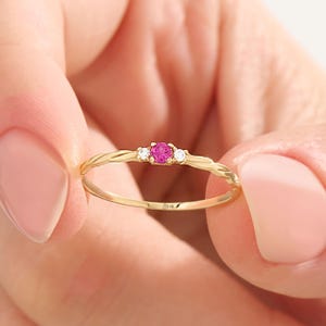 May include: A gold ring with a twisted band design, featuring a central pink gemstone flanked by two smaller white gemstones. The ring is held between two fingers, showcasing its delicate details and craftsmanship.