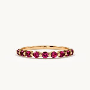 May include: A gold band ring featuring a series of round, vibrant pink gemstones set in a row. The metal band has a polished finish and includes a small, visible 14k stamp on the inner surface, showcasing a delicate and symmetrical design.
