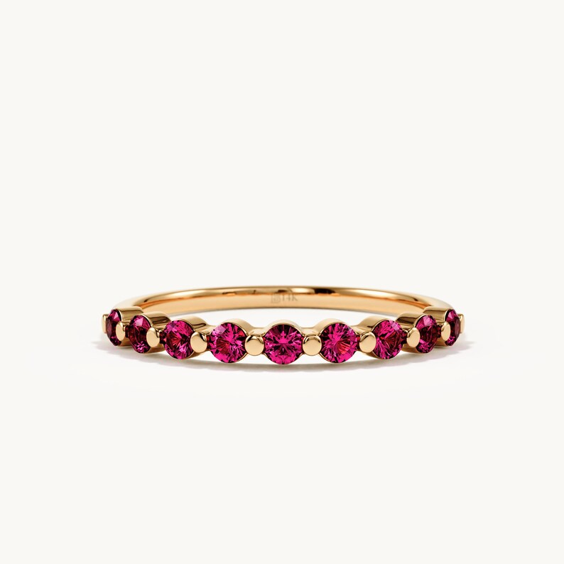 May include: A gold band ring featuring a series of round, vibrant pink gemstones set in a row. The metal band has a polished finish and includes a small, visible 14k stamp on the inner surface, showcasing a delicate and symmetrical design.