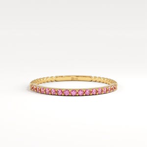 May include: A thin gold band ring featuring a row of small, round, bright pink gemstones set along the front half. The metal band has a textured, beaded edge design and is stamped with the text 14K on the inner surface of the ring.
