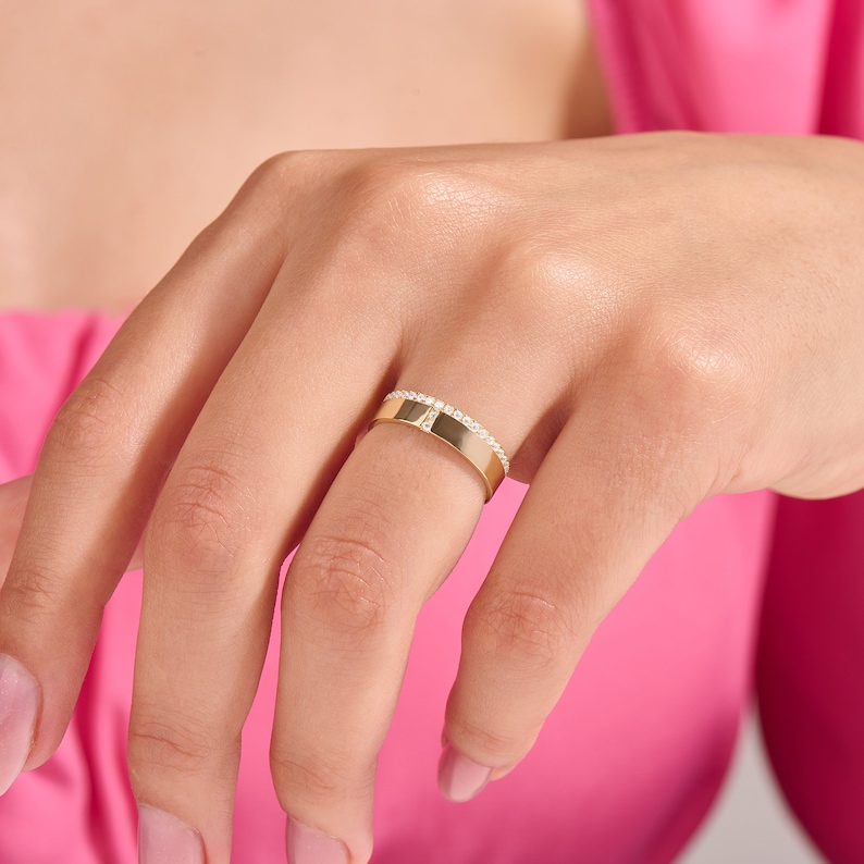 May include: A gold band ring with a row of small, clear stones set along one edge. The ring is on a finger, with a pink background.