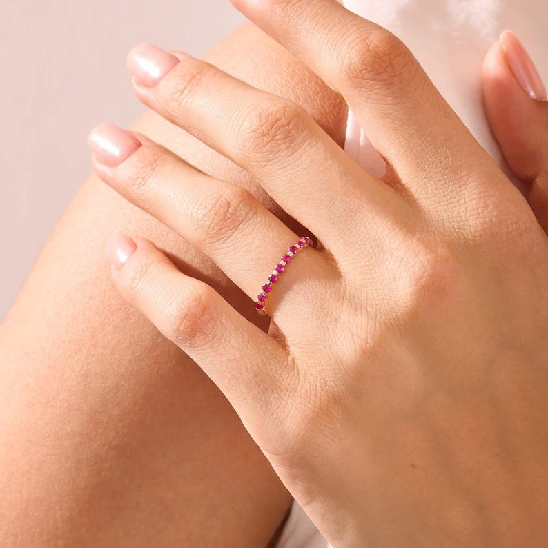 May include: A thin gold band ring featuring a continuous row of small, round, vibrant pink gemstones set along the top half of the band. The ring is worn on a finger, showcasing the delicate metal setting and the bright, saturated colour of the stones.