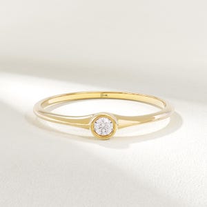 May include: A delicate gold ring with a single, round, clear gemstone set in the center. The ring band is thin and polished, with a subtle widening at the gemstone setting. The ring is marked with "14K".