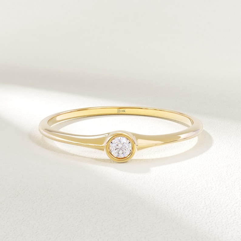 May include: A delicate gold ring with a single, round, clear gemstone set in the center. The ring band is thin and polished, with a subtle widening at the gemstone setting. The ring is marked with "14K".