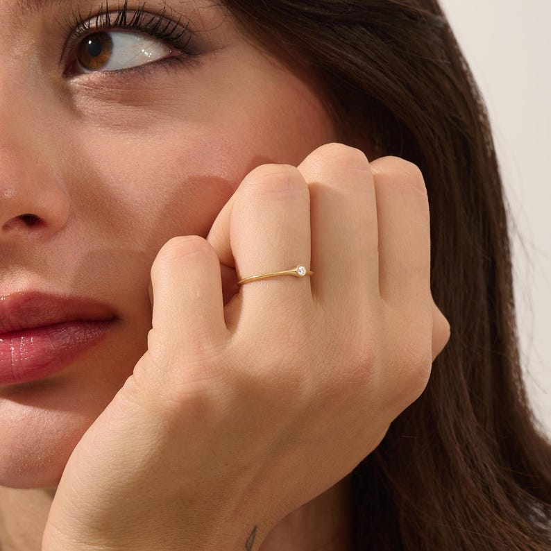 May include: A delicate gold ring with a small, round, clear gemstone. The ring is simple and elegant, with a thin band. The ring is on a person's finger.
