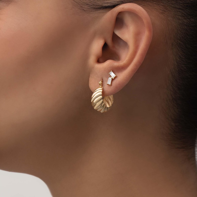 May include: A gold-toned hoop earring with a ribbed, textured surface and a separate stud earring featuring two small, clear, rectangular baguette-cut stones arranged in a vertical line, both worn on the earlobe.
