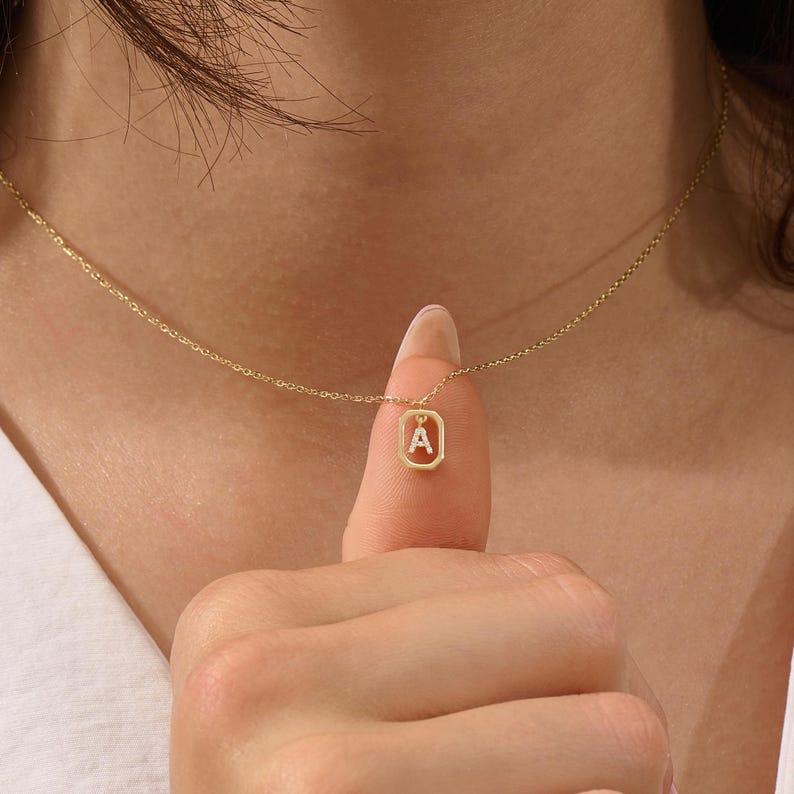 May include: A delicate gold necklace with a rectangular pendant displaying the letter "A". The pendant hangs from a fine chain. The necklace is a simple, elegant piece of jewellery.
