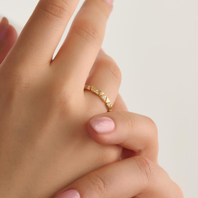 May include: A gold ring with a geometric design featuring small, triangular shapes. The ring is adorned with tiny, sparkling gemstones set within the triangles. The ring is worn on a finger, showcasing its intricate details and elegant style.