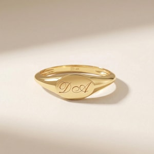 May include: A gold signet ring with an oval face. The ring is engraved with the initials "D A" in a delicate script. The ring is made of polished gold and has a smooth, rounded band. The ring is set against a neutral background.