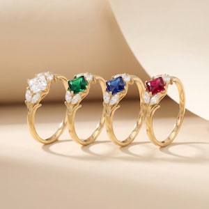 May include: Four gold rings with different colored square gemstones: white, green, blue, and red. Each ring features a leaf design with small white accent stones. The rings are displayed on a neutral background.