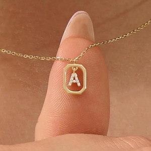 May include: A gold necklace featuring a delicate chain and a rectangular pendant frame. Inside the frame hangs a small, capital letter A charm encrusted with tiny, clear, sparkling stones. The piece is crafted from a polished yellow gold metal.