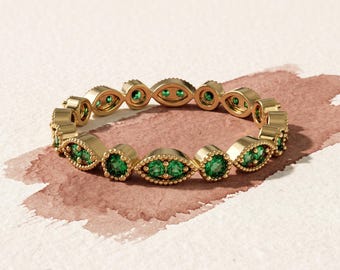 Puede incluir: Un anillo de oro con un patrón repetitivo de engastes en forma de marquesa, cada uno con dos pequeñas piedras verdes de corte redondo. La banda de metal tiene una textura de cuentas en los bordes, creando un diseño circular continuo.