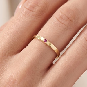 May include: A polished gold band ring featuring a single, small, round, vibrant pink gemstone set flush into the metal surface. The ring has a smooth, flat profile and a high-shine finish, showcasing a minimalist design with a central stone accent.