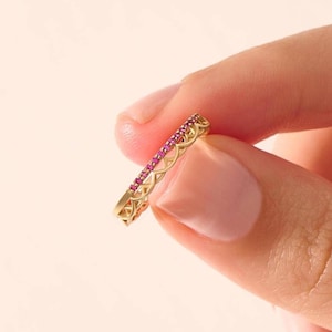 以下が含まれることがあります： 小さな明るいピンク色の宝石が並んだ、金色のリング。リングは、繰り返される幾何学模様の透かし彫りデザインです。リングは、柔らかいピンク色の背景に対して、2本の指の間に挟まれています。