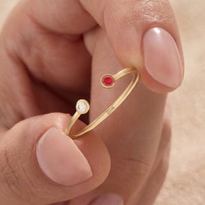 May include: A gold open-style ring featuring a small round red gemstone at one end and a small round clear white gemstone at the other. The band is stamped with the text 14K, indicating the gold purity of the jewelry piece.