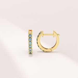 Puede incluir: Un par de pendientes de aro dorados con una fila de piedras preciosas de color azul claro. Los pendientes se muestran sobre un fondo blanco. Las piedras preciosas están espaciadas uniformemente a lo largo de la parte delantera de los aros.