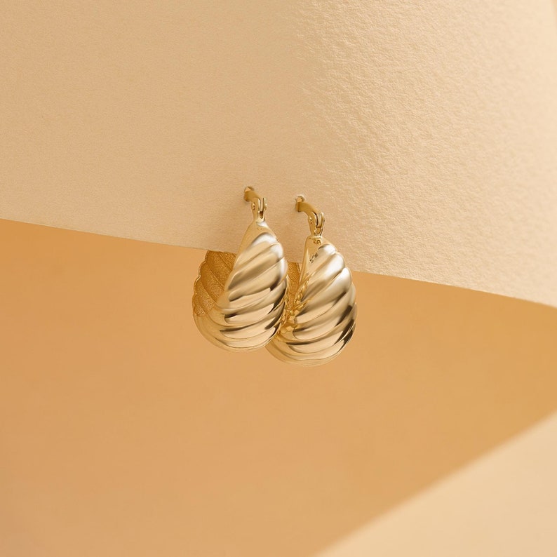 May include: A pair of gold hoop earrings featuring a thick, croissant-inspired ribbed texture. The earrings have a polished, reflective finish and a hinged post closure mechanism, showcasing a rounded, chunky silhouette with a curved design.