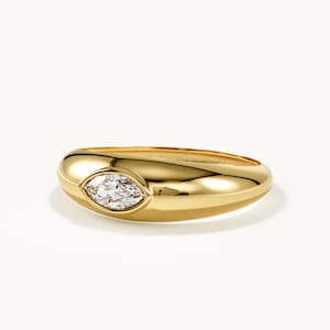 May include: A polished gold band ring featuring a single marquise-cut clear gemstone set horizontally in the center. The ring has a smooth, rounded, and thick profile that tapers slightly toward the back, showcasing a reflective metallic finish.