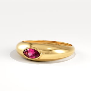 Könnte beinhalten: Ein goldener Ring mit poliertem Finish. Der Ring hat einen leuchtenden, oval geformten, rosa Edelstein in der Mitte. Das Band hat ein glattes, abgerundetes Design. Der Ring wird auf einer weißen Oberfläche präsentiert.