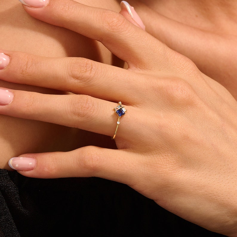 May include: A gold ring featuring a square, dark blue gemstone at the center, accented by smaller white gemstones. The ring has a delicate band and is displayed on a hand with painted nails.
