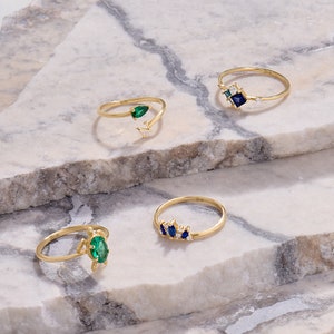 May include: Four gold rings with gemstone accents are displayed on a marble surface. The rings feature emerald and sapphire stones in various designs. One ring has an emerald teardrop shape, and another has a cluster of blue stones.