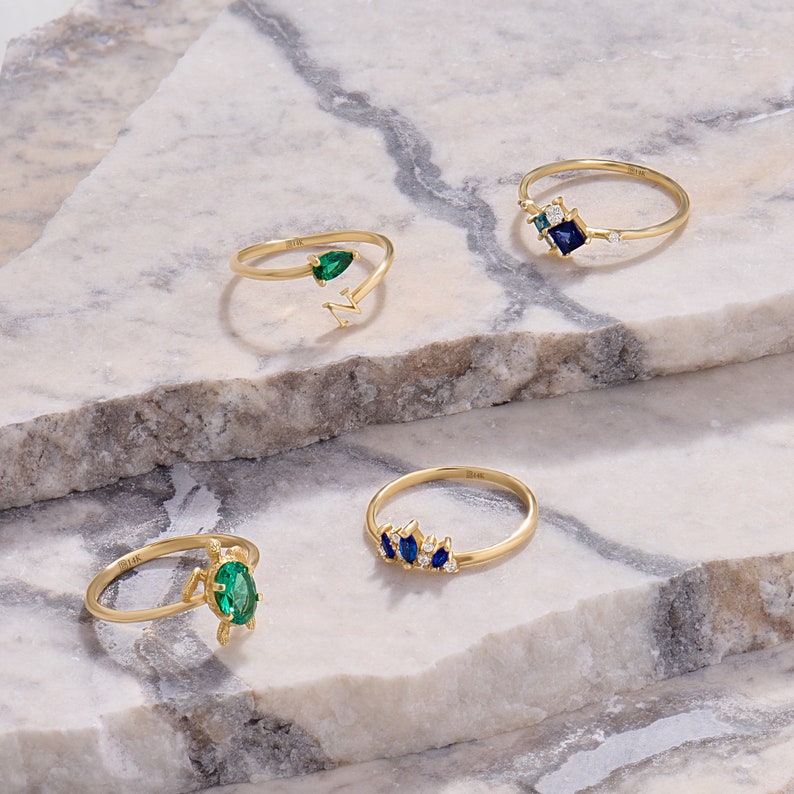 May include: Four gold rings with gemstone accents are displayed on a marble surface. The rings feature emerald and sapphire stones in various designs. One ring has an emerald teardrop shape, and another has a cluster of blue stones.