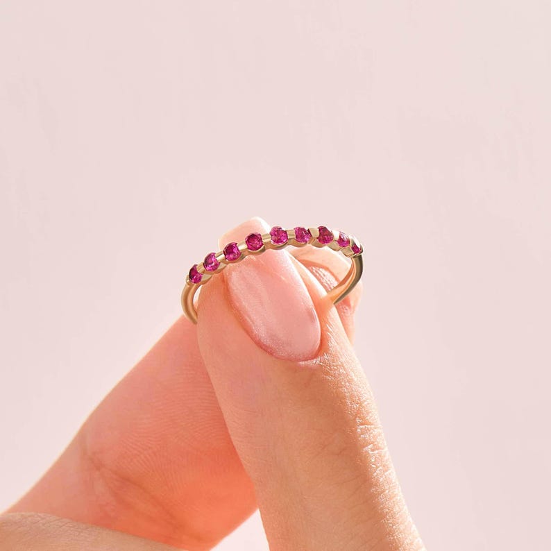 May include: A thin gold band ring featuring a row of nine small, round, vibrant pink gemstones set along the top half of the band. The ring is held between two fingers, showcasing the delicate metalwork and the bright, uniform colour of the stones.