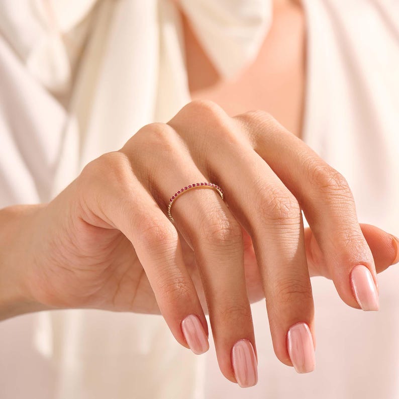 Puede incluir: Un anillo de oro con una banda de peque&ntilde;as piedras preciosas rojas. El anillo est&aacute; en un dedo, con un fondo suave y neutro. El dise&ntilde;o del anillo es delicado y elegante, adecuado para el uso diario o ocasiones especiales.