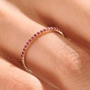Puede incluir: Un anillo de oro con una banda de peque&ntilde;as piedras preciosas rojas. El dise&ntilde;o del anillo es delicado, con las piedras preciosas muy juntas. El anillo se muestra sobre un fondo color piel, resaltando su color y dise&ntilde;o.